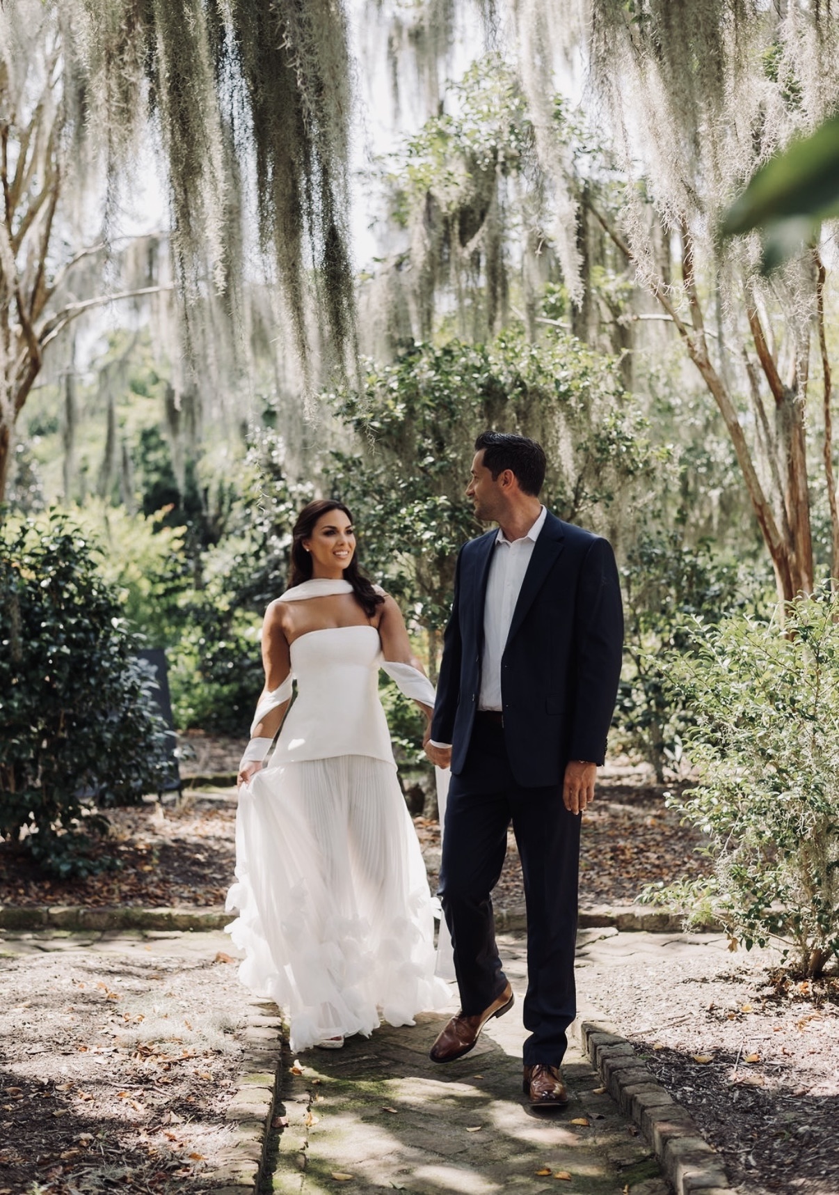 Chloe smiling toward Sam as they walk hand in hand beneath Spanish moss